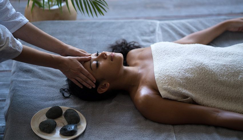 A woman wrapped in a towel lies on a massage bed as a pair of hands massage her temples.